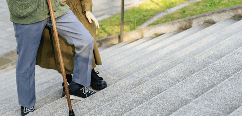 An older adult climbs stairs with a cane while a woman offers support to prevent a fall
